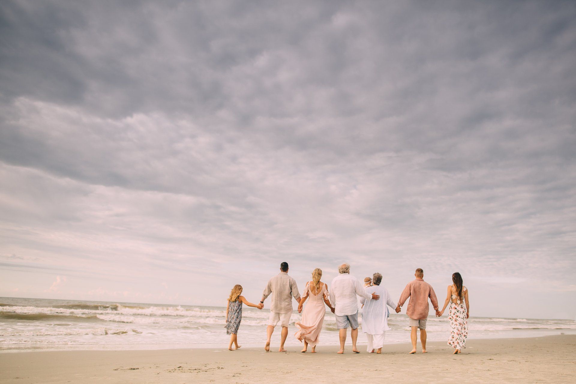 Family holding hands on beach, backs to camera, overcast sky.