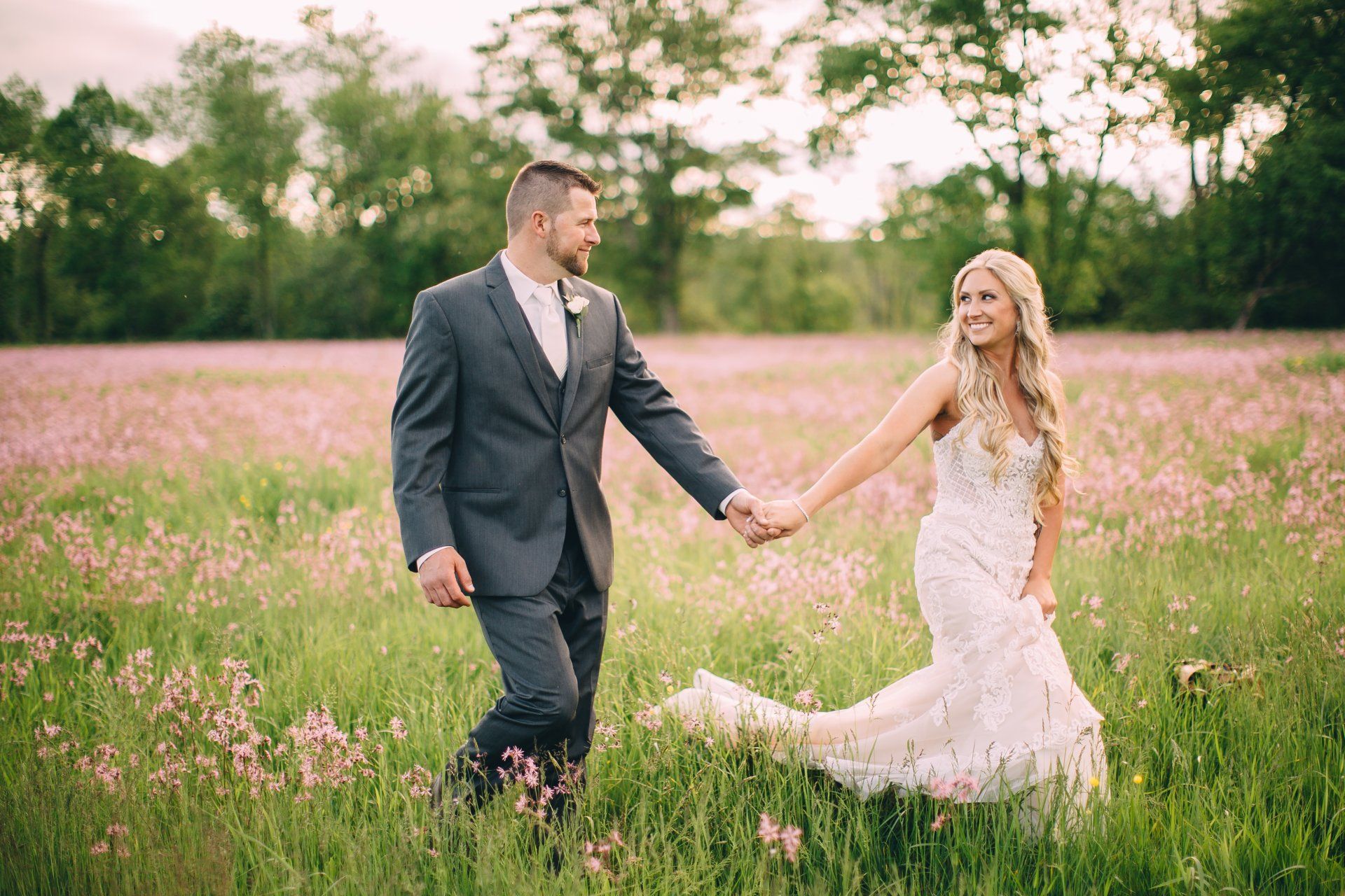 Bride and groom holding hands, walking in a field of pink flowers, groom in suit, bride in white dress.