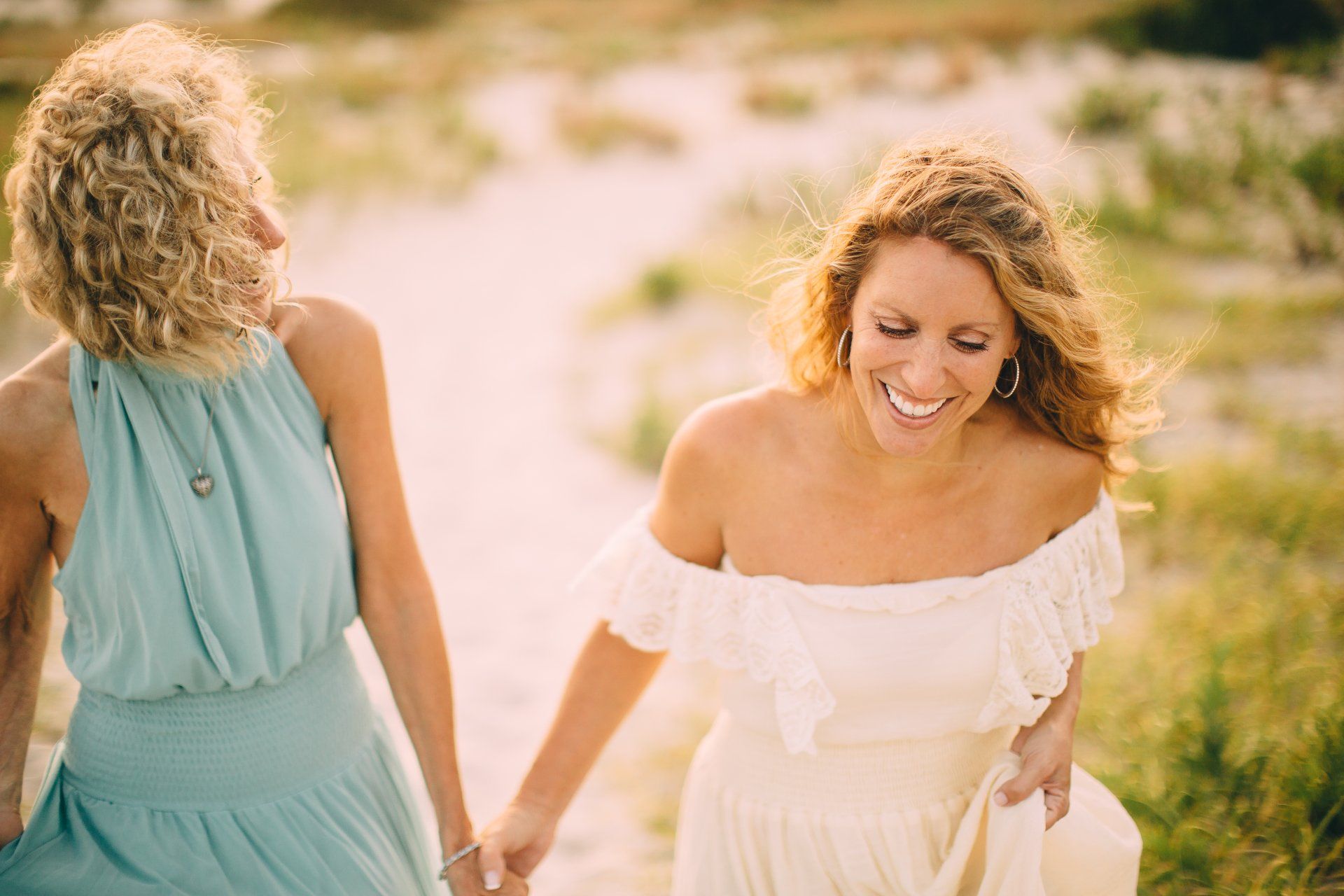Two women holding hands, walking on a sandy path, smiling. One wears a blue dress, the other a white off-the-shoulder dress.