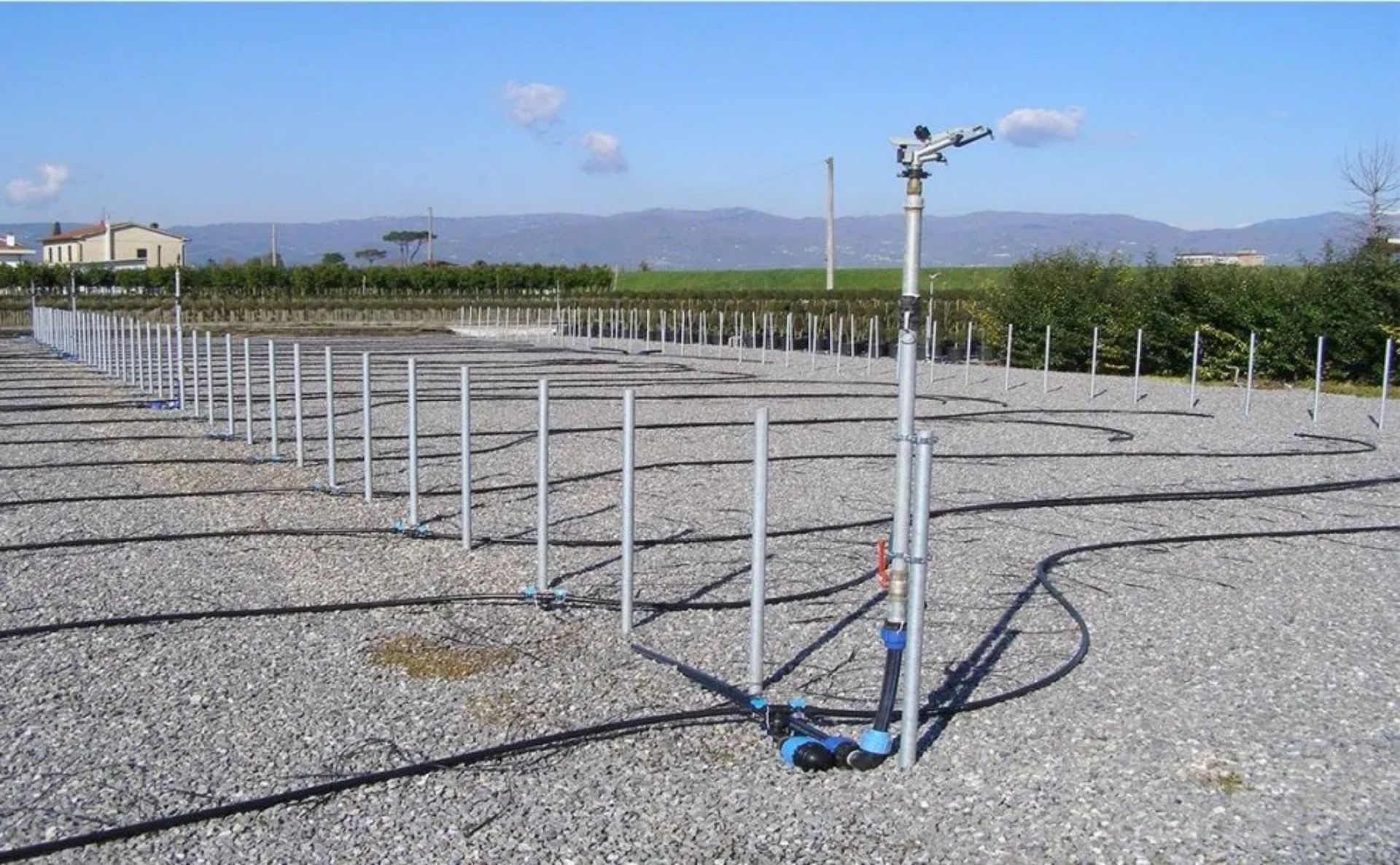 Sistema di irrigazione con tubi metallici e irrigatori su terreno ghiaioso, campi e montagne sullo sfondo.