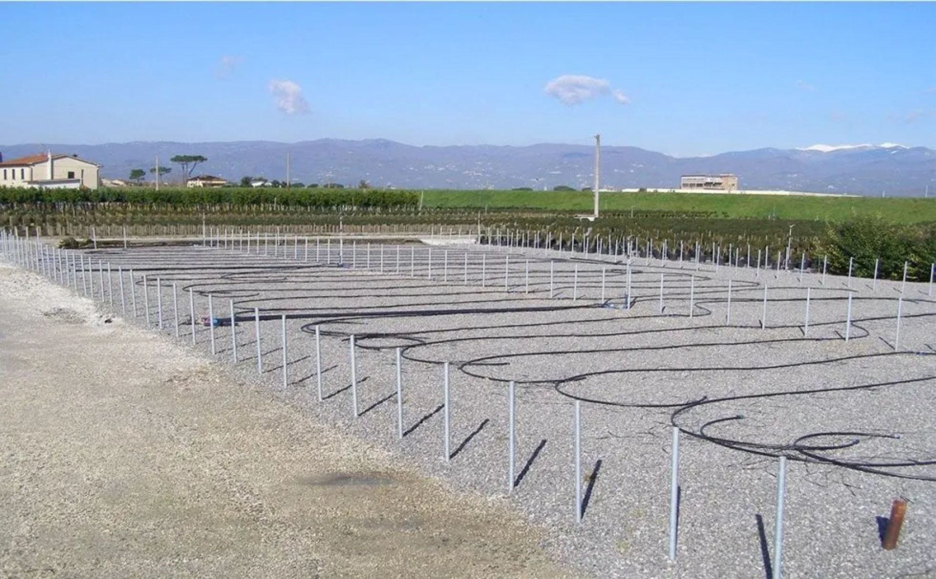 Terreno ghiaioso grigio con tubi e pali metallici, probabilmente per l'irrigazione, con sfondo rurale e montagne.