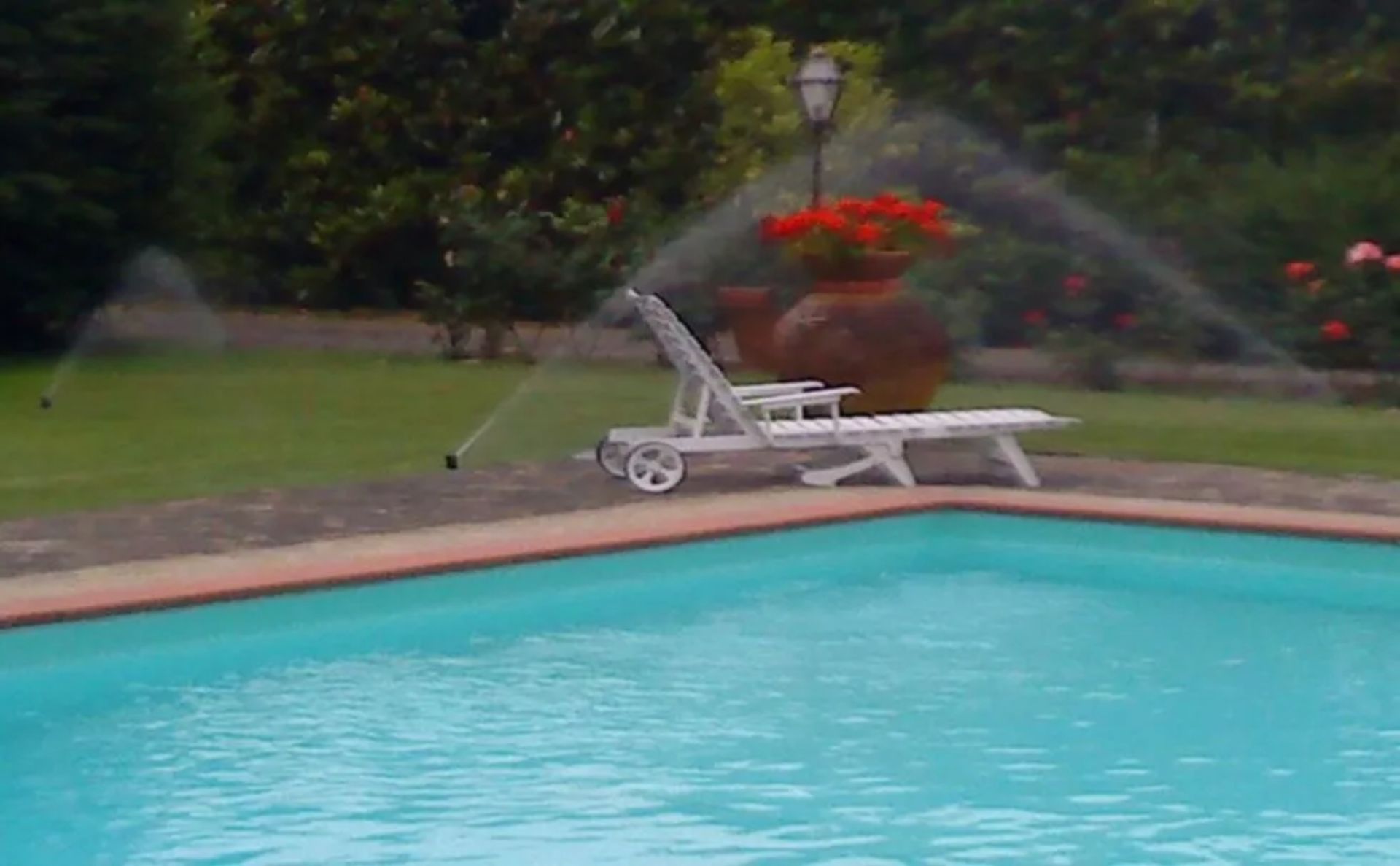 Irrigatori da giardino che spruzzano acqua vicino a una piscina con una sedia a sdraio e fiori rossi in vaso sullo sfondo.