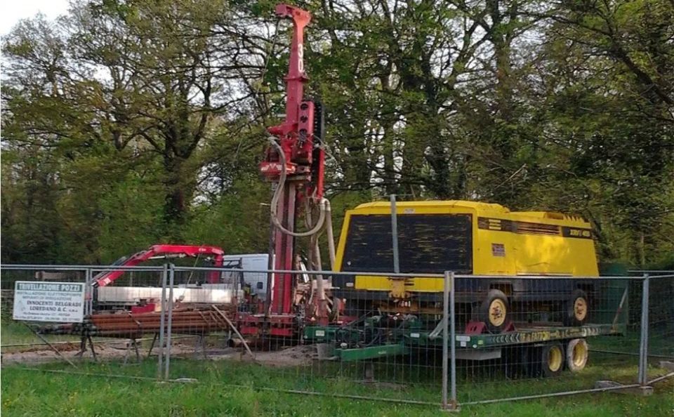 Impianto di perforazione su un rimorchio dietro una recinzione in una zona erbosa con alberi; attrezzatura gialla e rossa.