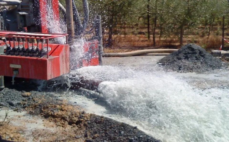 Acqua che sgorga da una macchina perforatrice, terreno fangoso, con un mucchio di terra sullo sfondo.