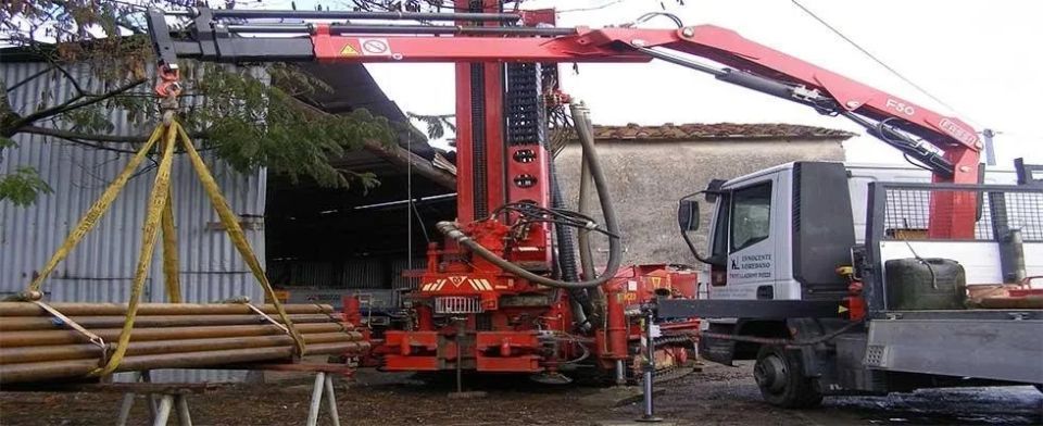 Una piattaforma di perforazione rossa solleva tubi. Un camion è parcheggiato vicino a un edificio.