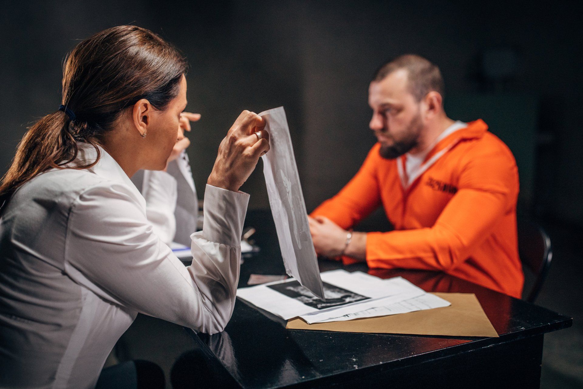 A woman is sitting at a table talking to a man in an orange jumpsuit.