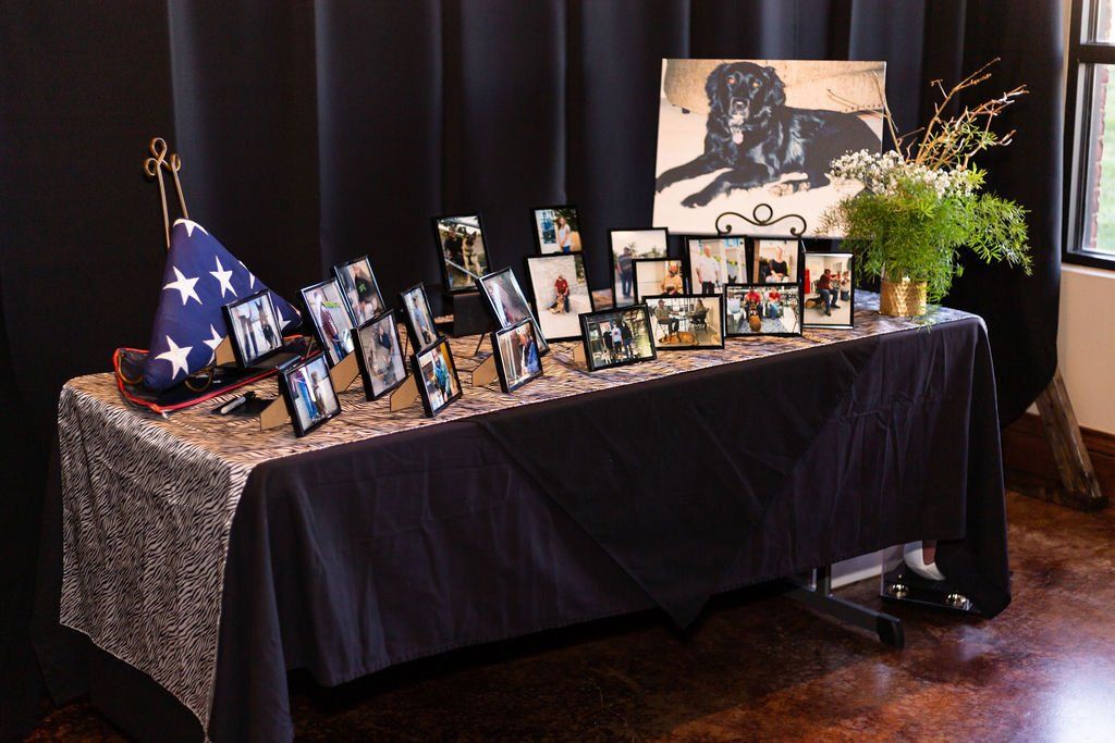 A memorial table covered in a black cloth holds a folded American flag, numerous framed photos, and a potted plant.
