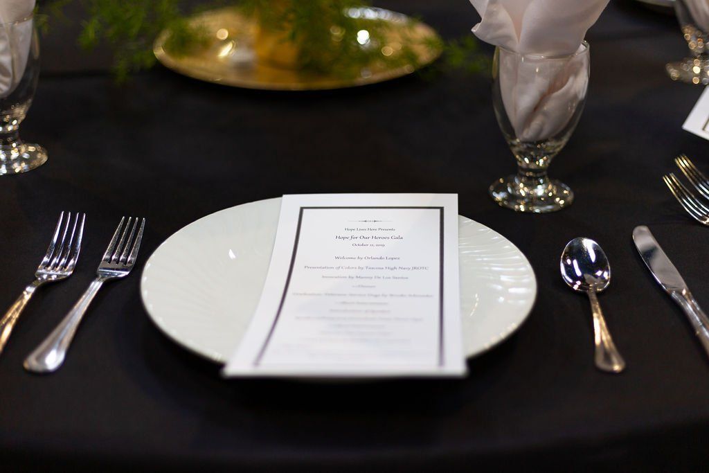 A formal table setting with a white plate, a printed menu, silver cutlery, and a water glass on a black tablecloth.