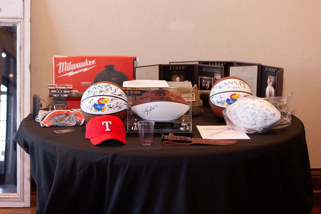 A black-covered table features autographed basketballs, a football, a red Texas Rangers hat, and a Milwaukee tool box.
