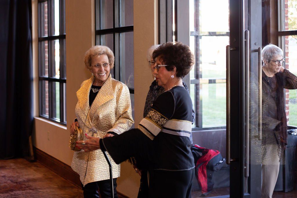 Three people stand in a sunlit room with tall windows, two conversing while a third person stands near the doorway.