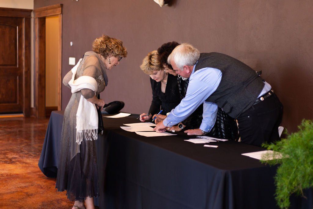 Three people stand behind a black-clothed table in a room with dark walls, reviewing documents while one person observes.