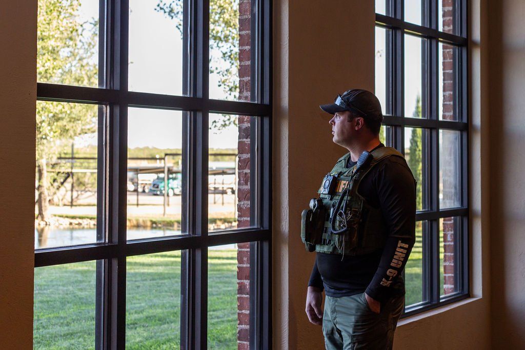 A person in a tactical vest and cap stands inside by a large window, looking out at a grassy area and trees.
