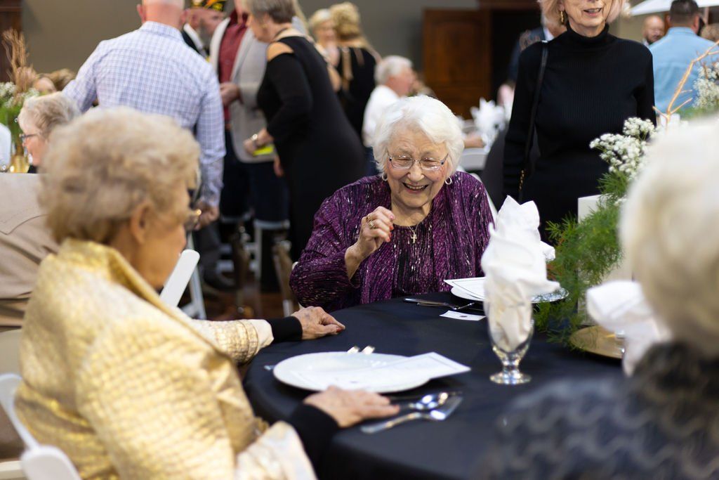 People seated at round tables at a social event, including a laughing person in a purple shirt and another in gold.