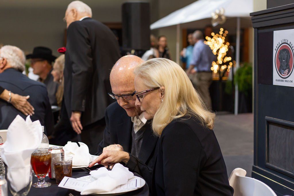 Two people in formal attire sit at a table, looking down at a paper while gesturing during an indoor event.