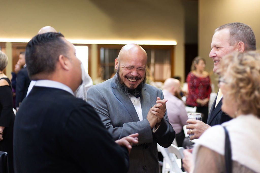 People in formal attire gather at an indoor event, with one person in a grey suit laughing while others talk nearby.