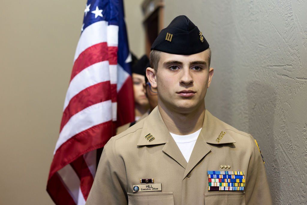 A service member in a tan uniform and garrison cap stands solemnly beside a vertical American flag.