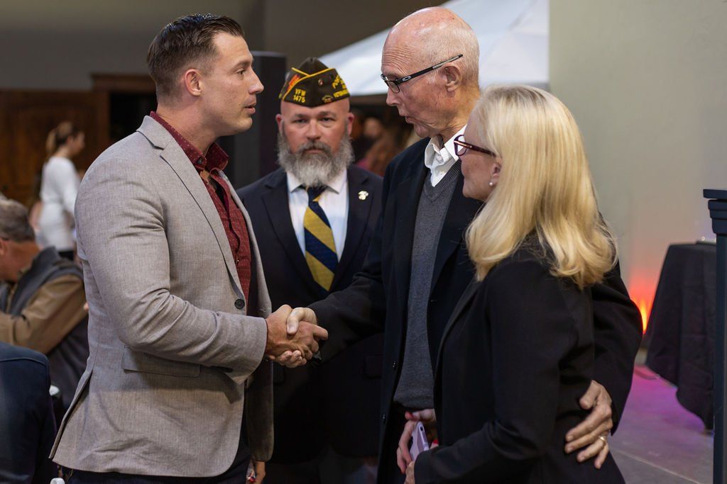 Four people in formal attire greet one another in a handshake at a public event.