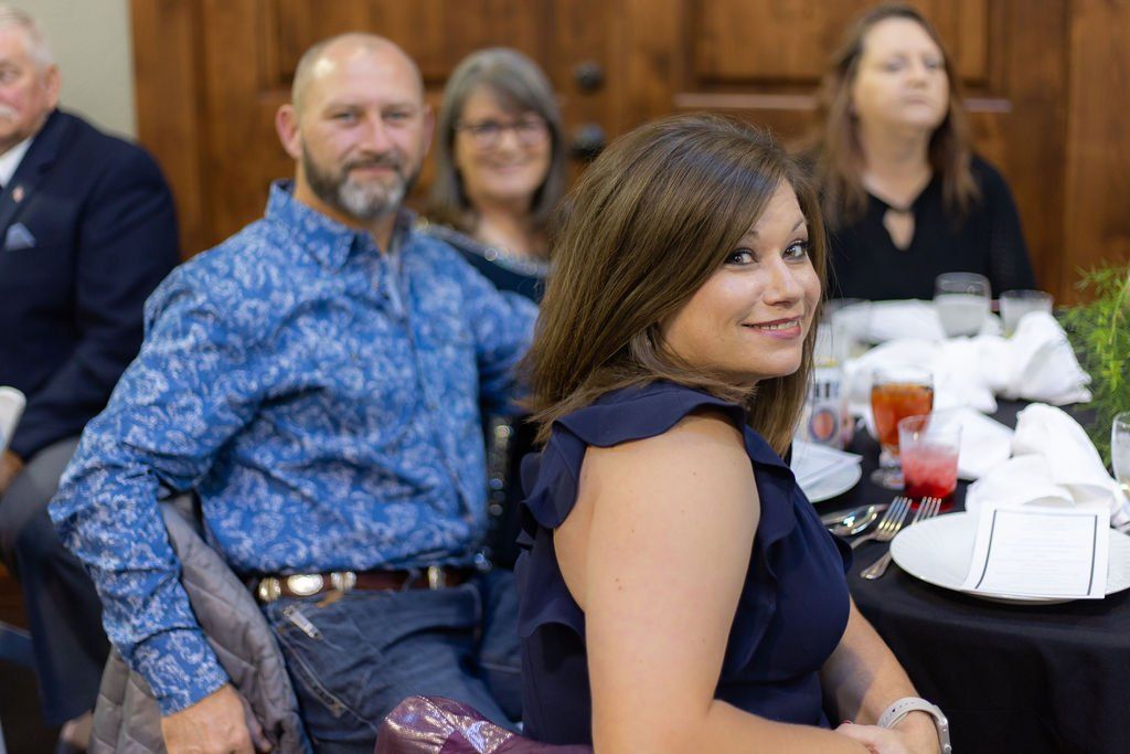 A group of people seated around a table at an event, with a woman in a navy dress in the foreground smiling at the camera.