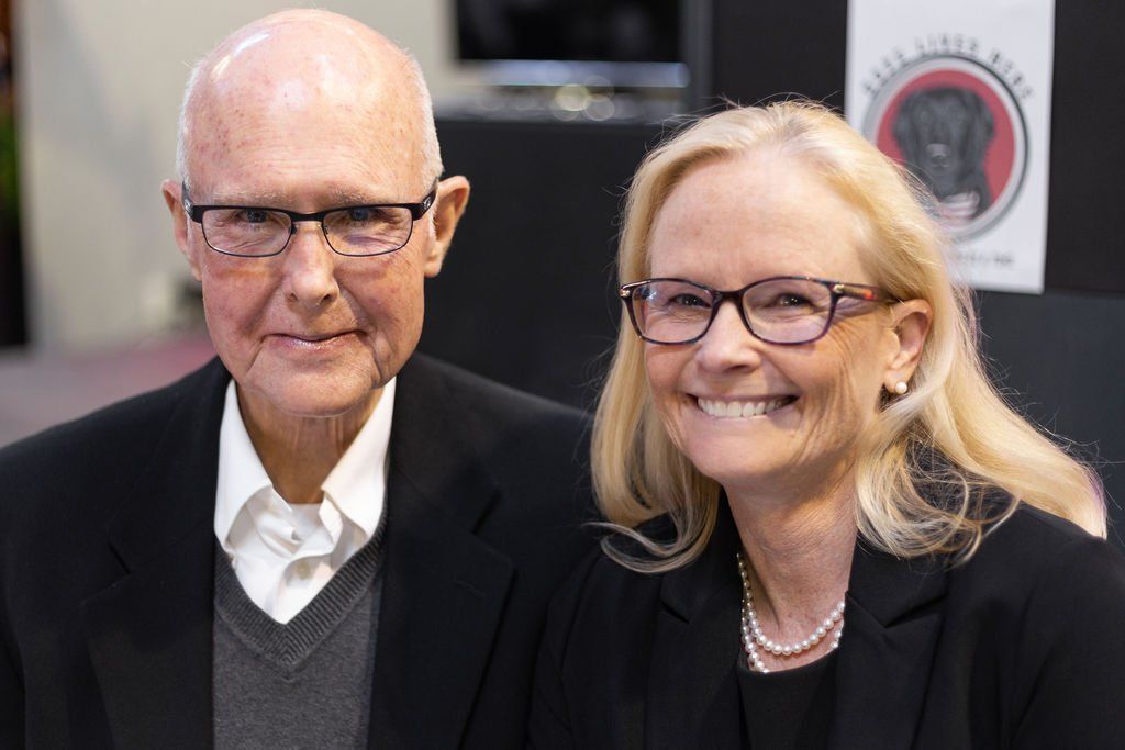 An older couple wearing glasses and professional attire smiles together in front of a sign with a dog emblem.