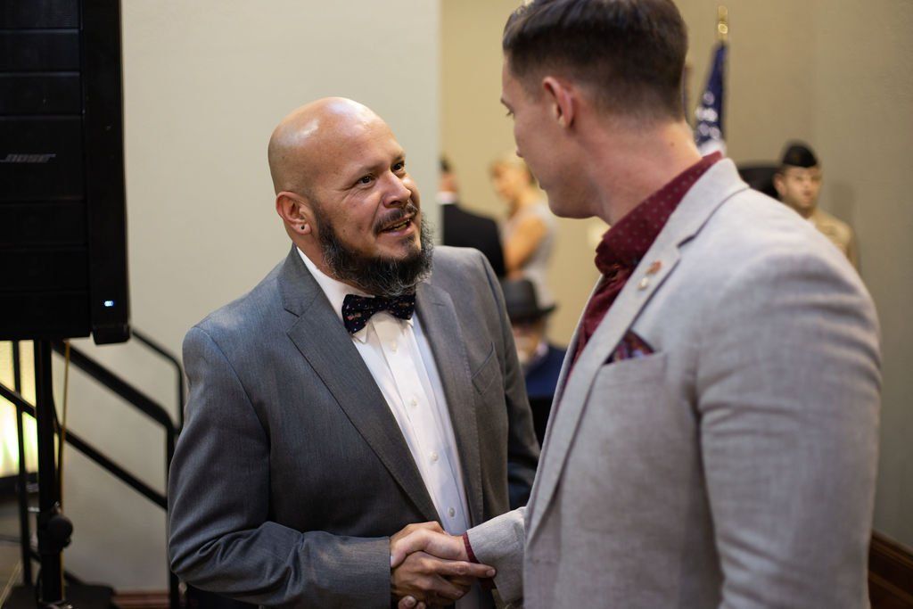 Two men in suits shaking hands while conversing at an indoor event.