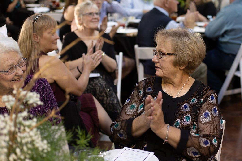 People seated at round tables in a decorated event space clap, smiling and looking toward a stage or presenter.