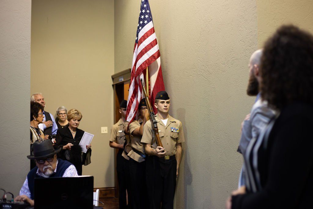 A color guard carries the American flag into a room as people watch, while a man in a hat sits at a computer nearby.