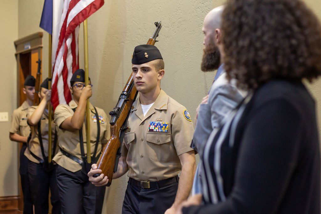 A color guard in tan uniforms carrying an American flag and a rifle walks past people standing in a room.