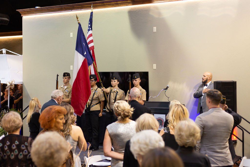 A color guard stands on a stage holding the US and Texas flags, facing a seated audience in a bright indoor room.