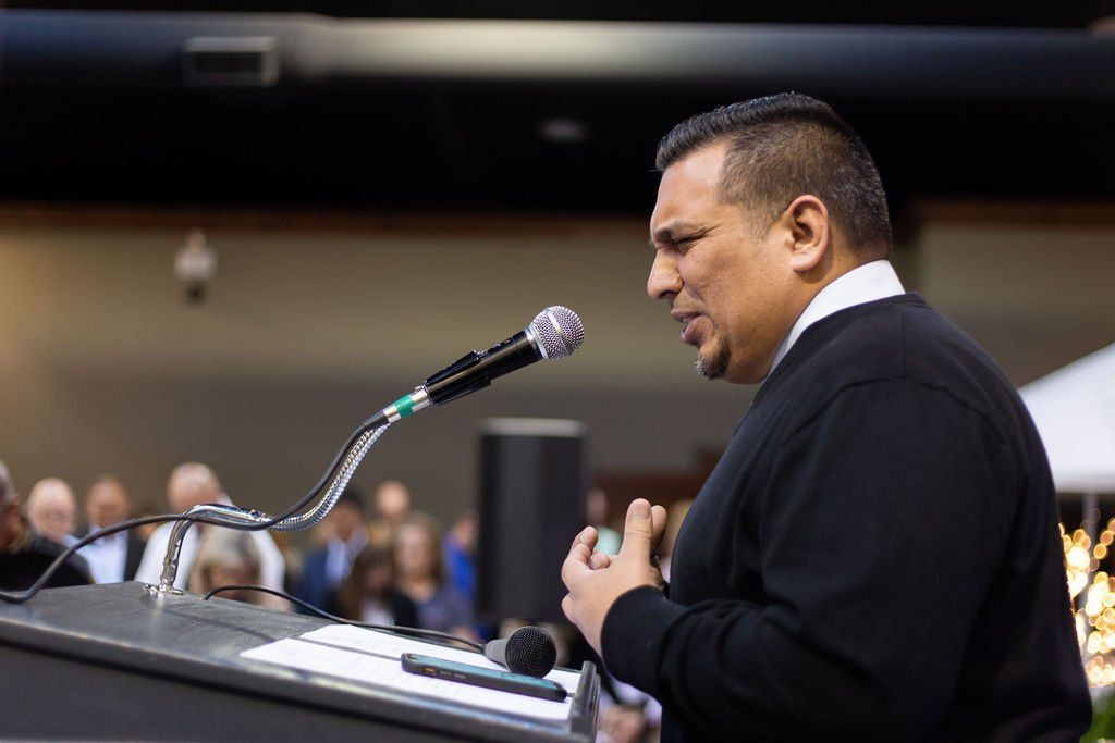 A person wearing a black sweater speaks into a microphone at a podium before a blurred audience.