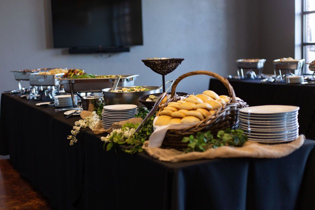 A catering buffet table with food warmers and a basket of bread rolls, set against a dark wall in a dimly lit room.