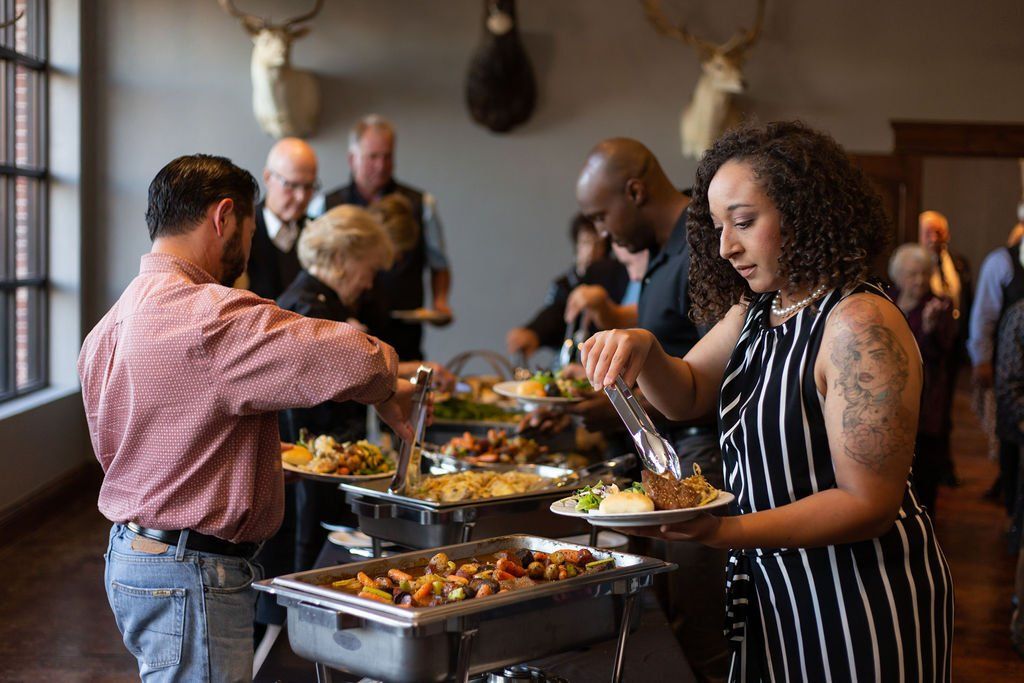People serve themselves from a buffet line featuring metal chafing dishes in a room decorated with animal mounts.