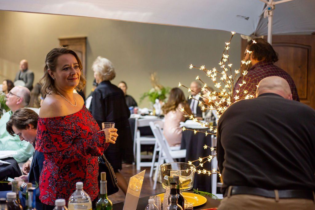A woman in a red patterned off-the-shoulder top holds a drink at a dimly lit indoor event with guests in the background.