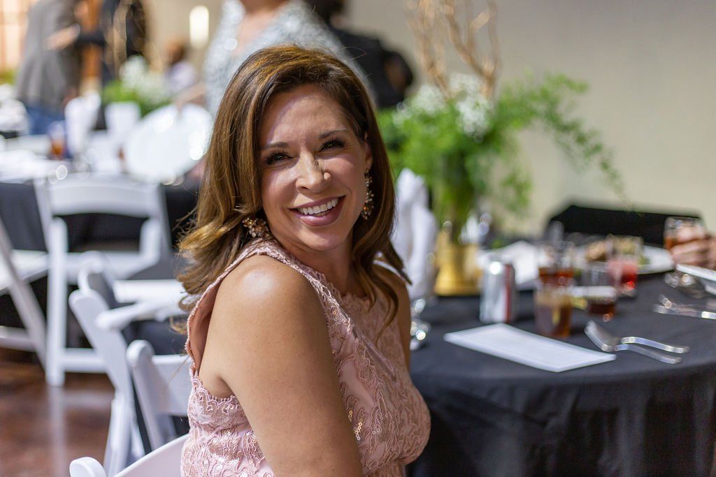 A smiling person in a sleeveless, textured light pink dress sits at a table in a formal event setting.