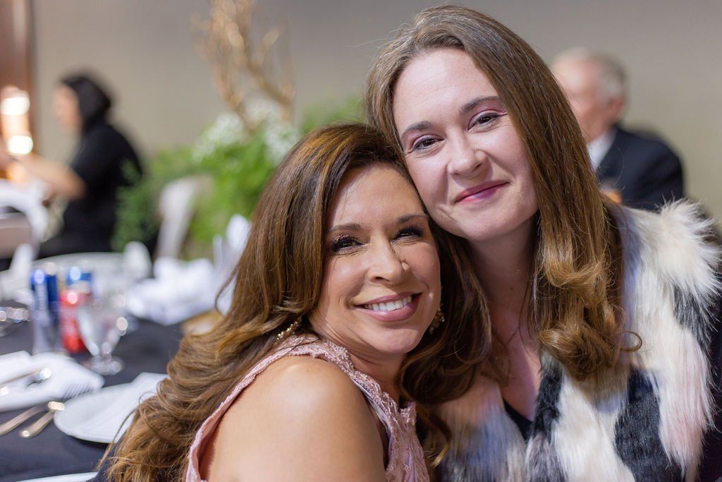 Two people with long brown hair lean their heads together and smile at the camera in a warmly lit indoor setting.