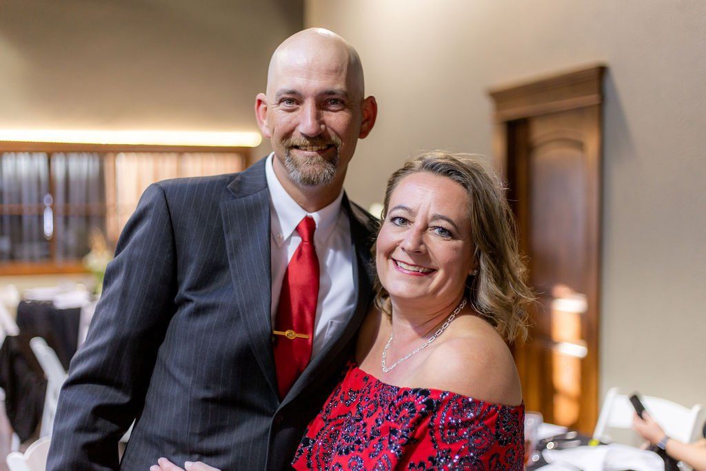 A person in a dark suit with a red tie and a person in a red patterned dress smiling together in a formal indoor setting.