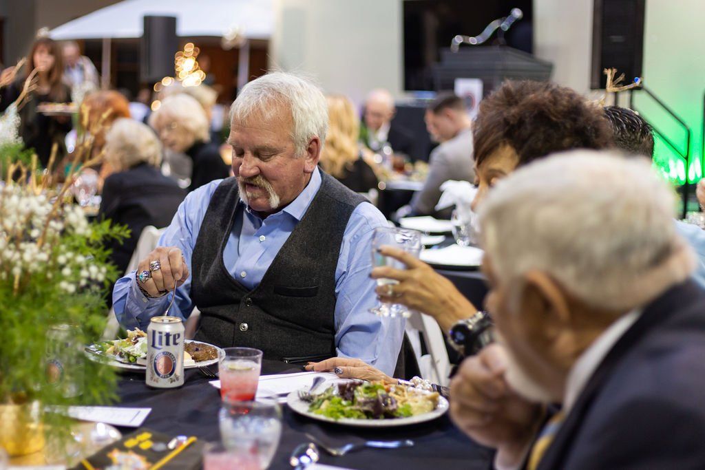 People dining at a formal event, featuring a man in a vest at a table with a Miller Lite can and plates of food.