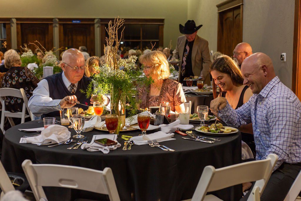 People dine at a round table with a black tablecloth and floral centerpiece in a formal hall.