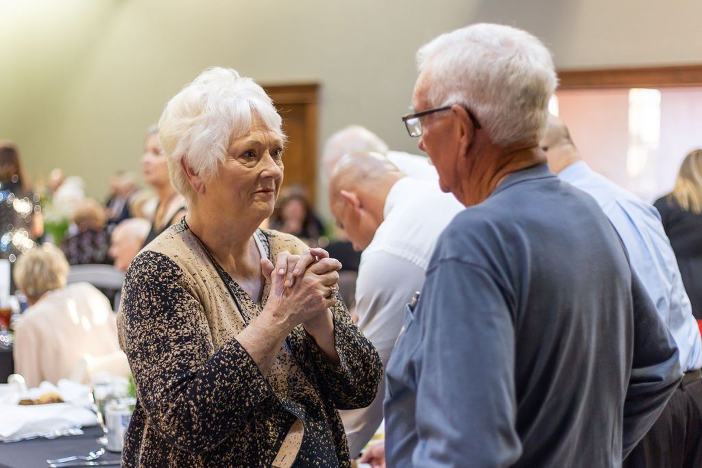 A person with short white hair talks to a person wearing glasses and a blue shirt at an indoor gathering.