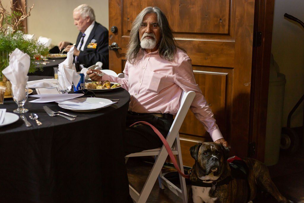 A person with long gray hair and a beard sits at a table in a dimly lit room with a brindle dog resting by their side.