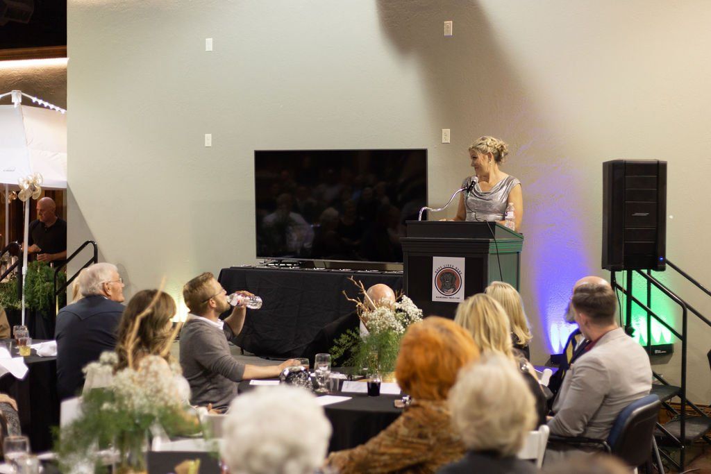 A person stands at a podium giving a presentation to an audience seated at tables in a large event space.