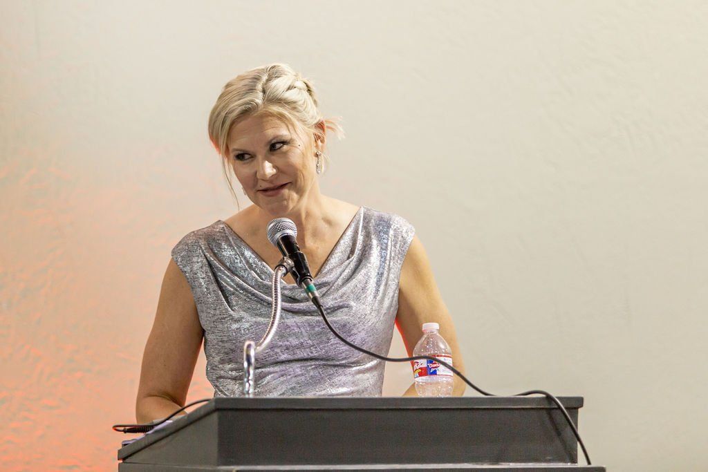 A person in a silver dress stands at a podium, speaking into a microphone, with a water bottle nearby.