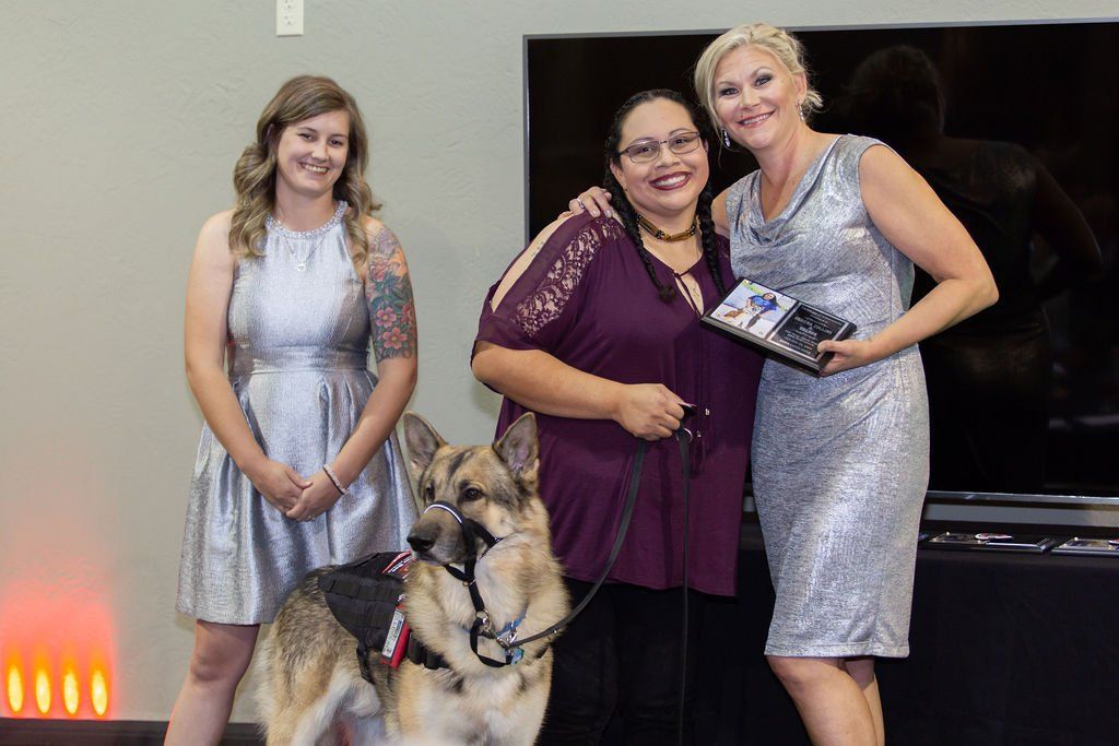 Three women stand together with a German Shepherd service dog, with two women in metallic silver dresses and one in purple.