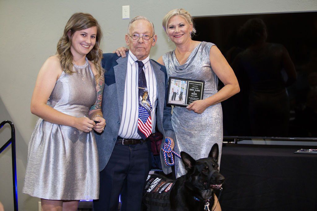 Two women in metallic silver dresses stand flanking a man wearing a suit, tie, and a service dog in a vest.