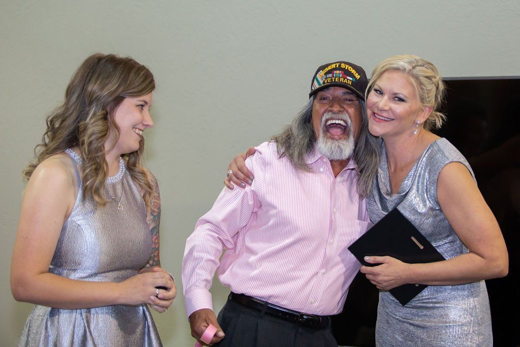 Two people in silver dresses stand on either side of a man in a pink shirt and veteran cap, all smiling and posing.