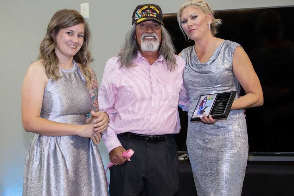 A person standing between two others in sparkling dresses, one of whom holds a small plaque, in an indoor setting.