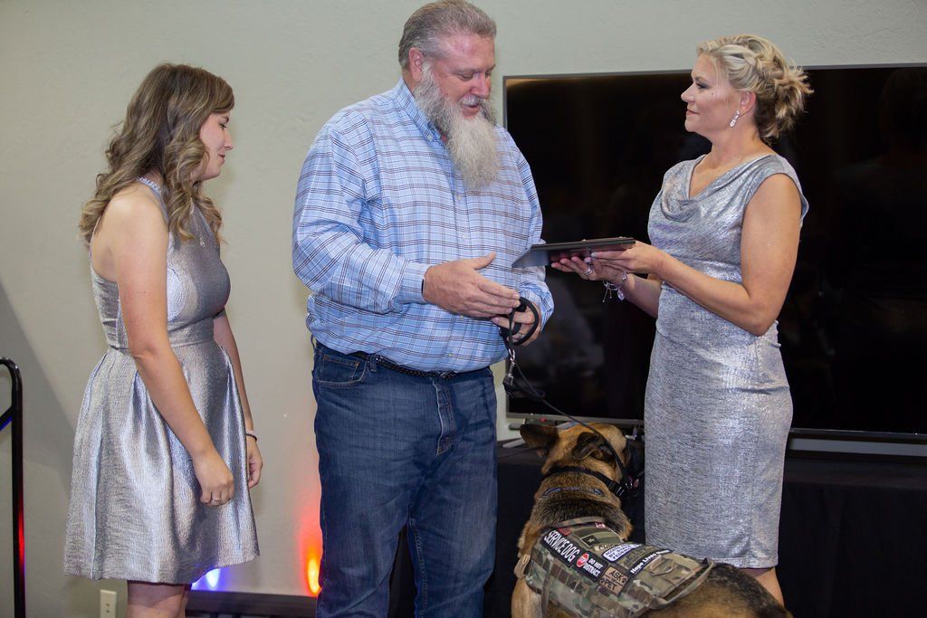 A man in a plaid shirt and blue jeans receives a plaque from a person in a silver dress, with another person watching.