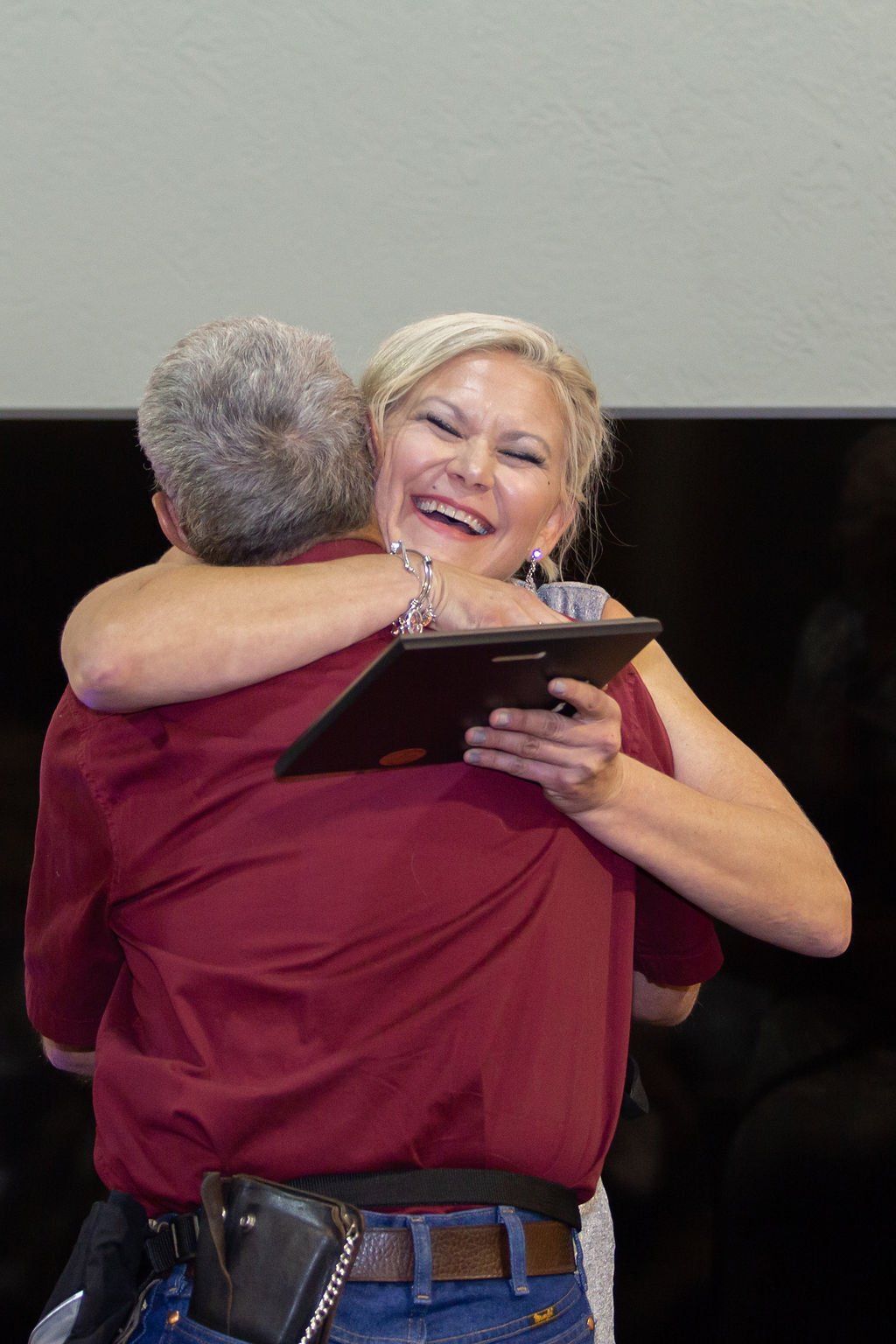 A person in a maroon shirt and blue jeans hugs a smiling person holding a framed award or certificate.