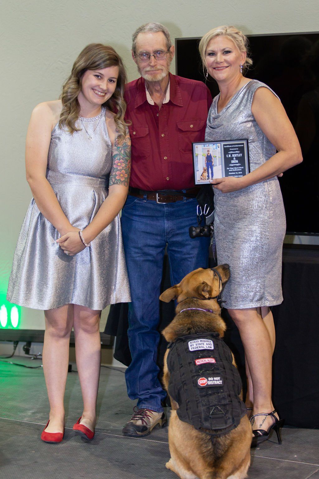 Two people in silver dresses and a man in a maroon shirt pose with a service dog wearing a tactical vest.