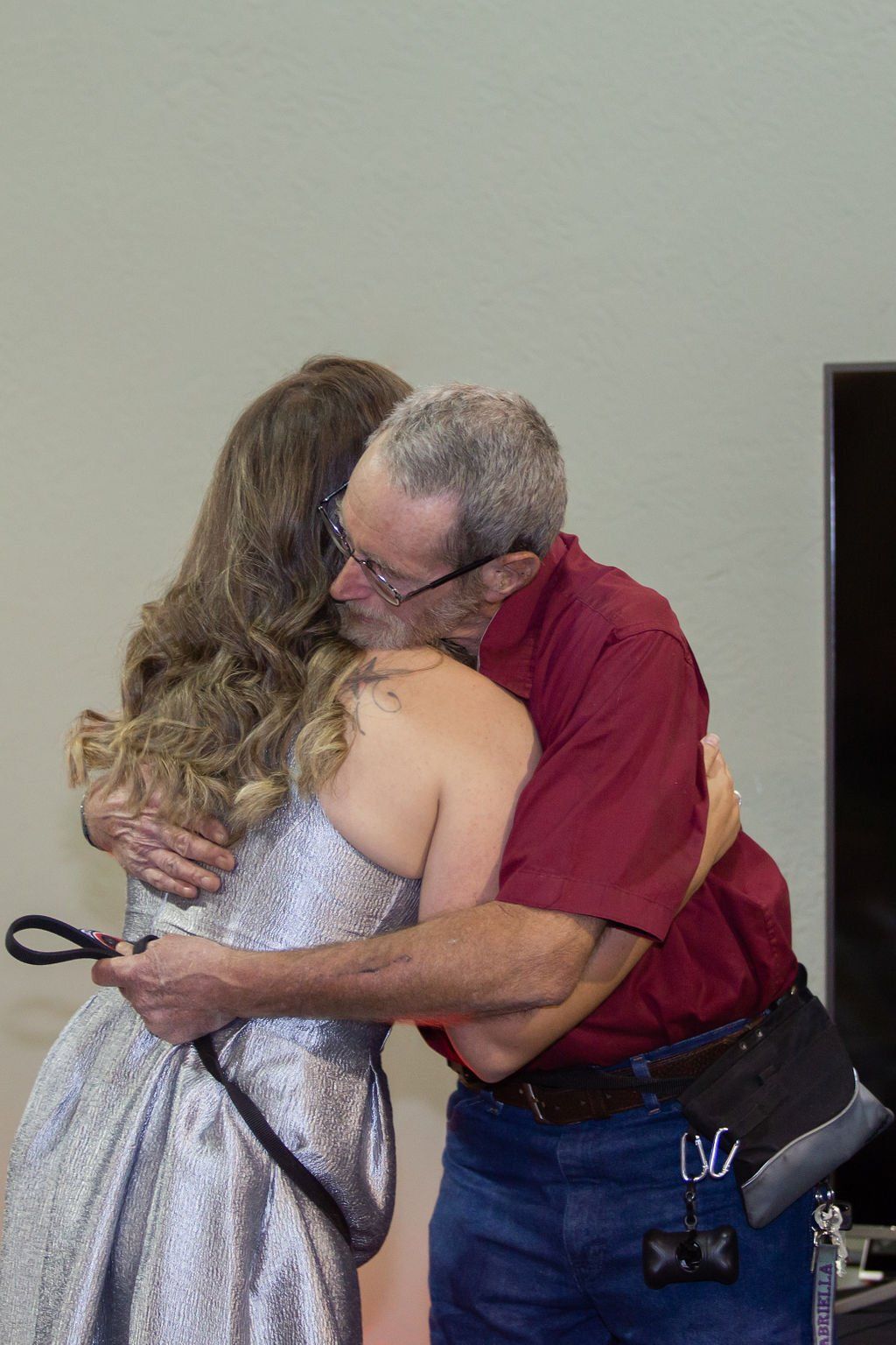 A person in a red shirt hugs someone in a formal silver dress, holding a black leash behind their back.