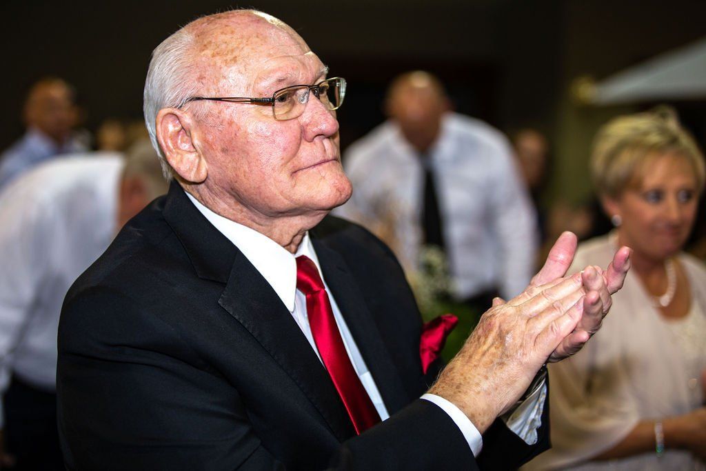 An elderly person in a suit and red tie applauds in a brightly lit, formal social setting.